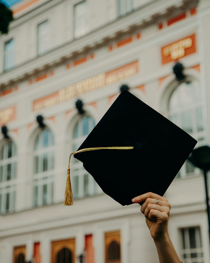 A graduate holding a mortarboard up in front of a university building