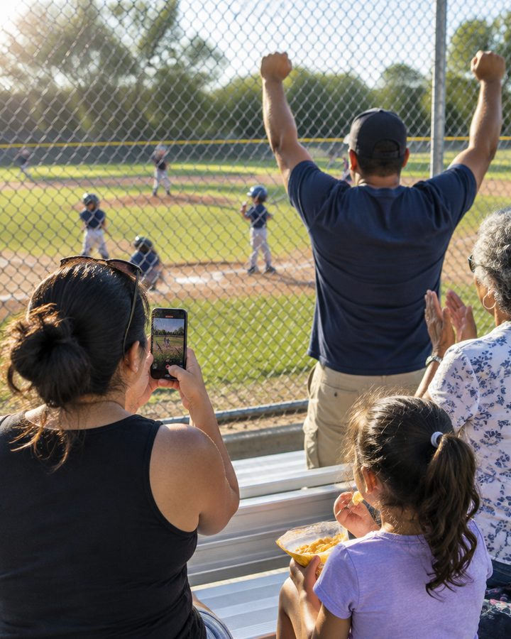 Parents in the bleachers at a youth baseball game, one filming and one cheering